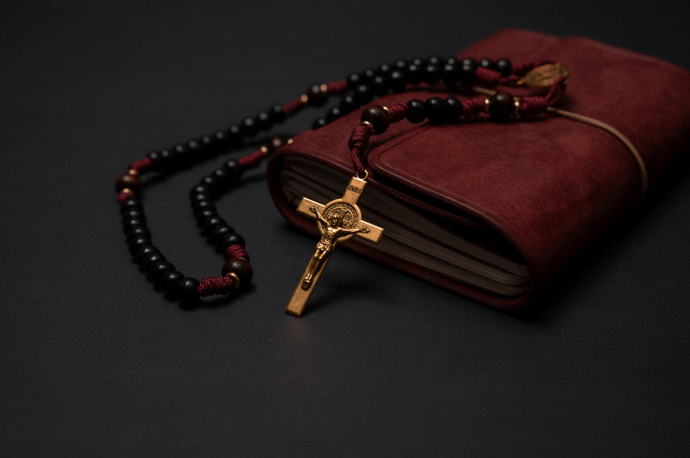 Handmade St. Benedict paracord rosary in burgundy cord with black wood beads, draped over a leather journal