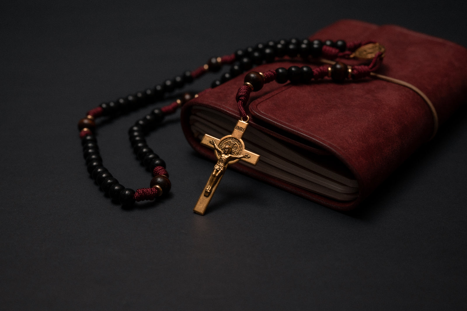 Handmade St. Benedict paracord rosary in burgundy cord with black wood beads, draped over a leather journal
